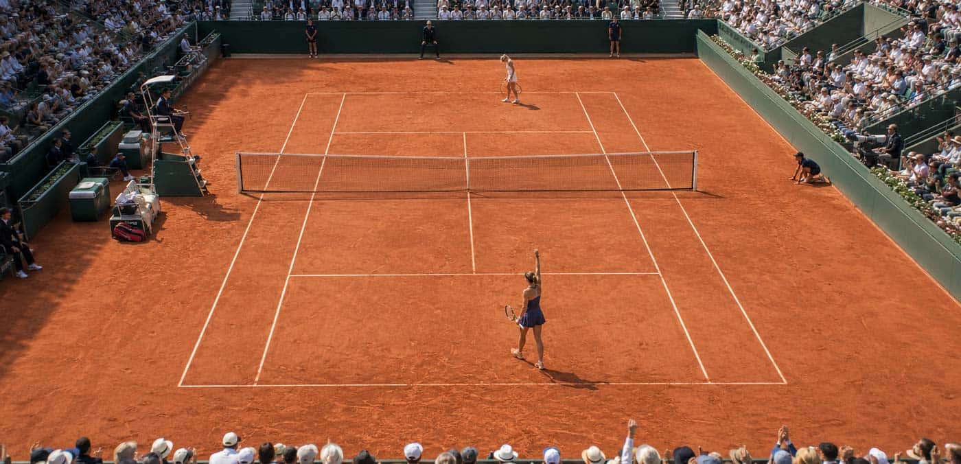 Vue panoramique en hauteur d’un court de tennis en terre battue avec une joueuse célébrant un point et son adversaire au fond du court.