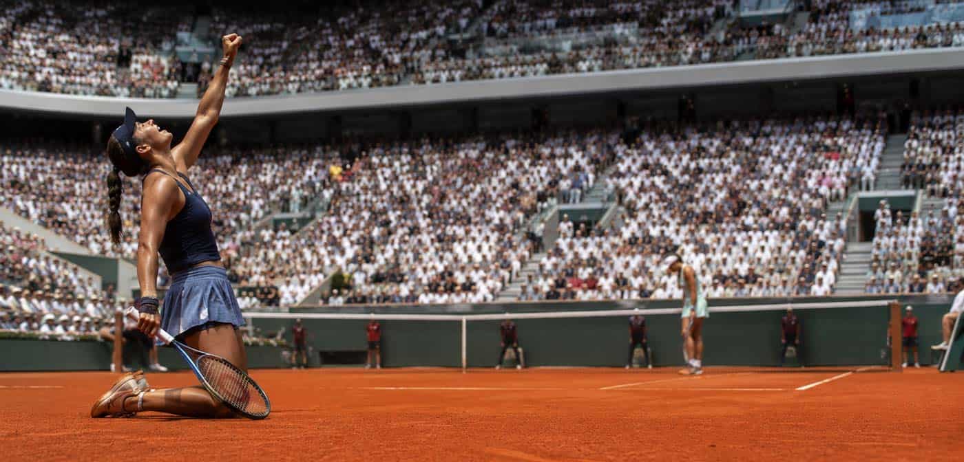 Joueuse de tennis à genoux sur terre battue levant le poing, avec son adversaire au fond du court dans un grand stade.